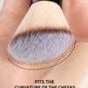 A close-up of a fluffy makeup brush demonstrating its curve, designed to fit the curvature of the cheeks for facial grooming.