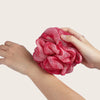 A hand is holding a vibrant pink exfoliating mesh bath sponge.