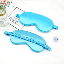 Dos máscaras para los ojos de seda azul claro exhibidas sobre un fondo blanco con un frasco de vidrio y un pequeño paquete.