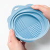 A hand holds a light blue silicone makeup brush cleaner and drying stand set with textured surfaces.