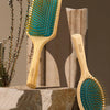 A wooden hair brush with teal bristles is displayed next to a plant on a stone surface.