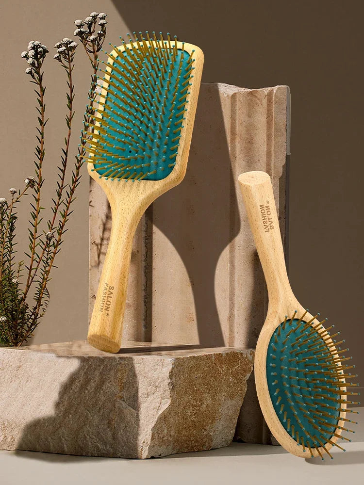 A wooden hair brush with teal bristles is displayed next to a plant on a stone surface.
