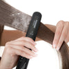 Une main utilise un fer à lisser professionnel en céramique noire sur des cheveux bruns.