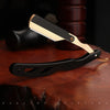 A professional straight edge barber razor with a black handle and gold accents resting on a wooden surface.