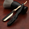 A black and gold professional straight edge barber razor lying on a dark wooden surface next to a grooming brush.