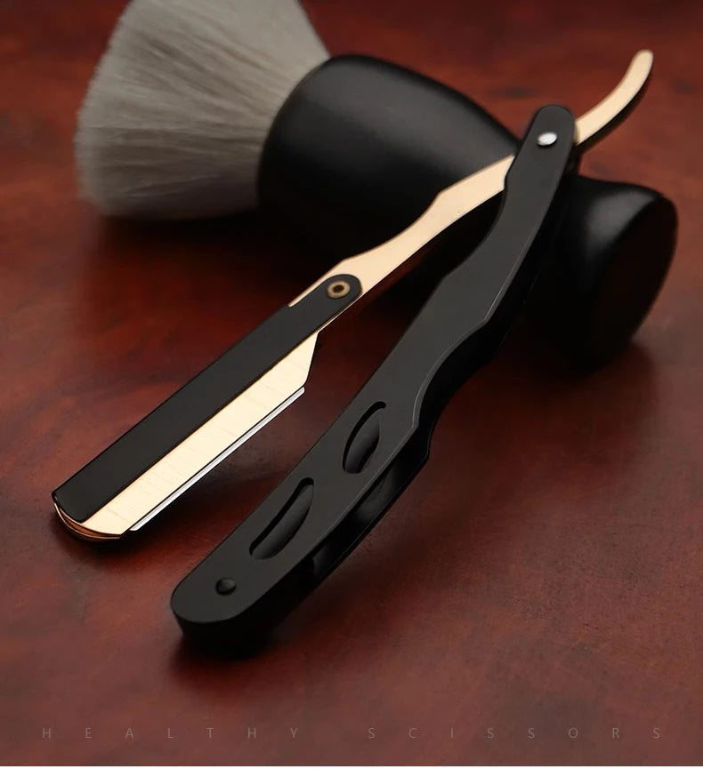 A black and gold professional straight edge barber razor lying on a dark wooden surface next to a grooming brush.
