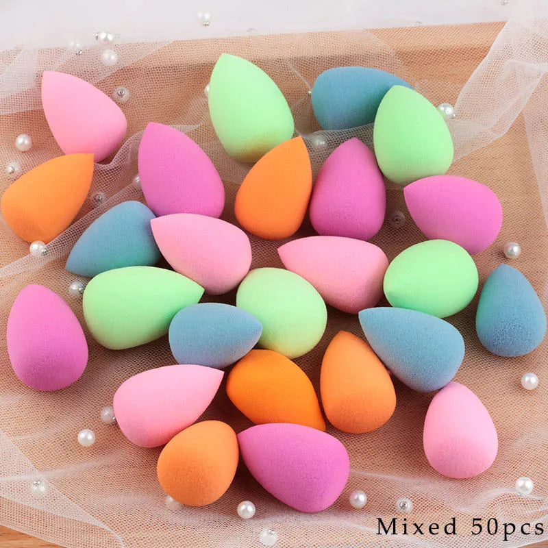A collection of 50 mixed colored makeup sponge puffs in various shades of pink, orange, and green arranged on a sheer fabric.