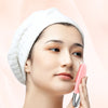 A woman is using a pink sonic facial cleansing brush on her face while wearing a white towel.