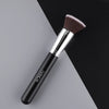 A black flat top kabuki foundation brush with a silver ferrule and bristles resting on a gray surface.