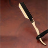A professional straight edge barber razor with a light wood handle and black blade rests on a dark brown surface.