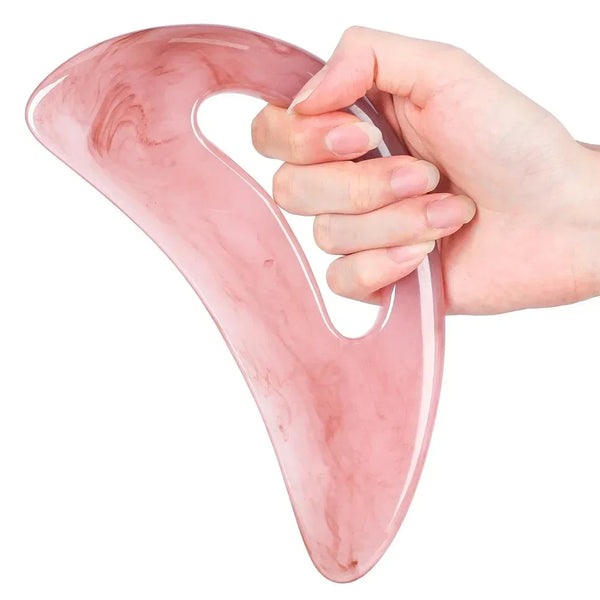 A hand holds a pink resin gua sha tool with marbled patterns for facial and body massage.