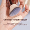 A hand holds a flat head foundation brush with soft gray and beige bristles for makeup application.