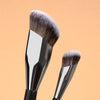 A professional makeup brush set featuring a large, dense brush and a smaller tapered brush against a light orange background.