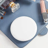 A round white LED makeup mirror resting on a blue-gray surface with cosmetic bottles nearby.