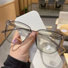 Una mano sostiene unas gafas cuadradas vintage y transparentes contra una superficie blanca.