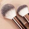 Un par de brochas de maquillaje profesionales con cerdas plateadas y blancas sobre un fondo claro.