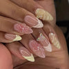A close-up of a hand displaying 24-piece almond press-on nails featuring delicate floral and butterfly designs in soft pink a