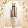 A white automatic ceramic hair curler stands in a soft-lit studio setting with floral accents.