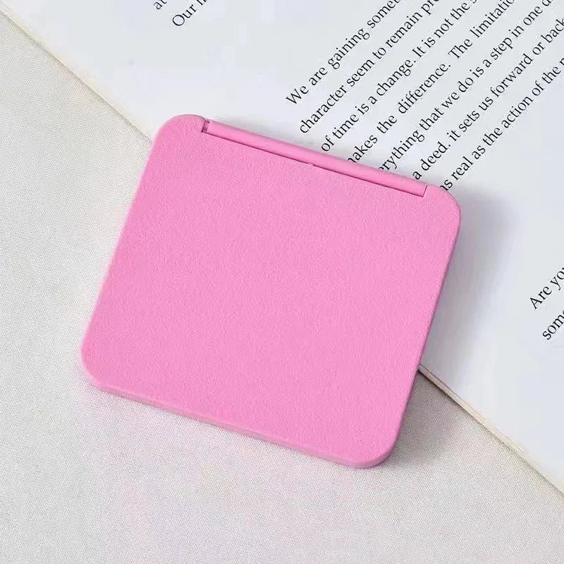 A pink double-sided portable makeup mirror lying on a white surface next to a book.