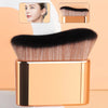 A close-up of a precision hand-held foundation makeup brush with black and gold accents, shown alongside a model's face.