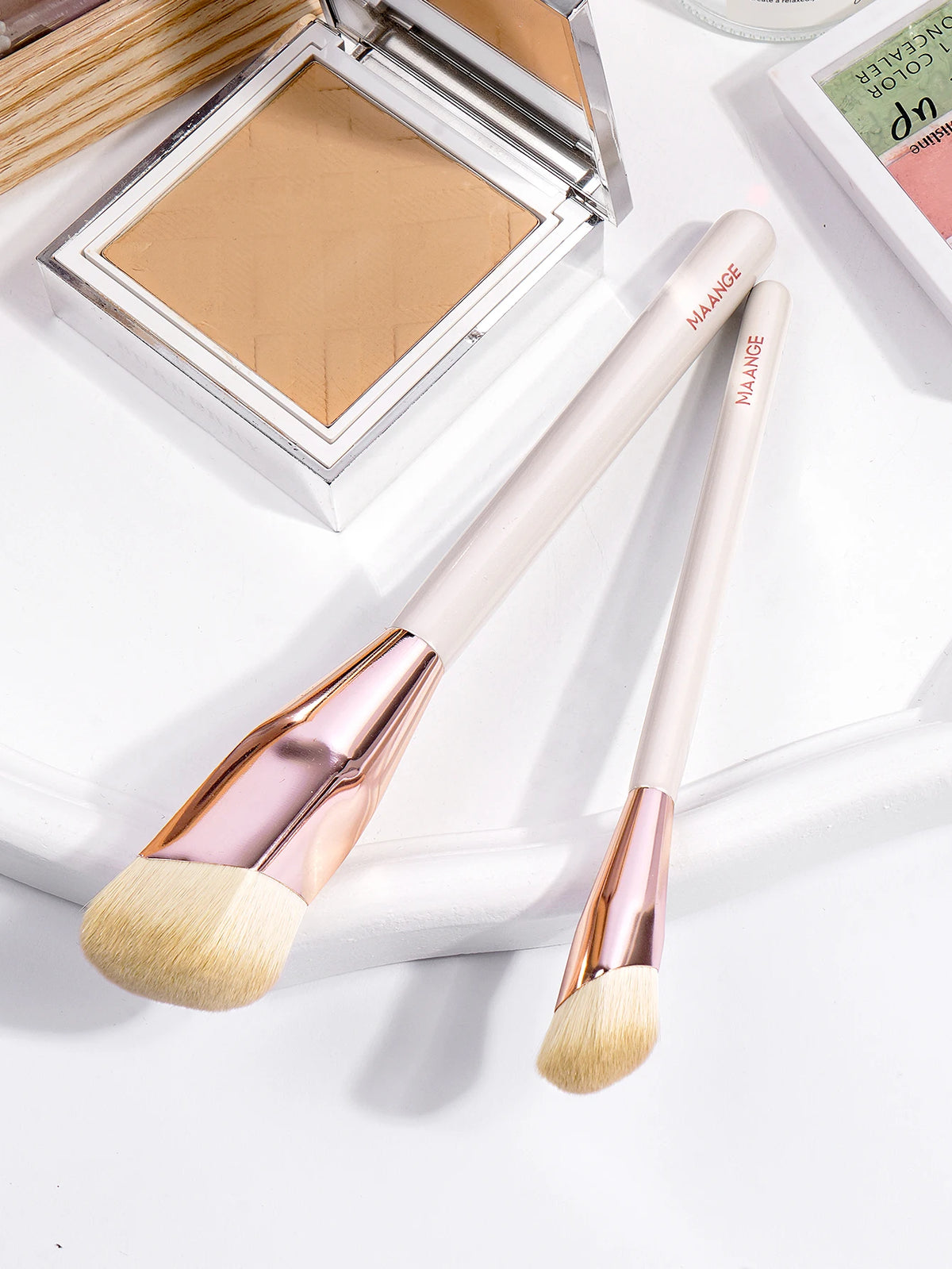 A pair of light pink makeup brushes with gold accents are displayed next to an open makeup palette.