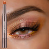 A cosmetic beauty glazed eyeshadow stick in a silver casing with a bronze-pink shade shown next to an eye.