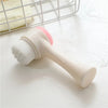 A beige facial brush with a pink silicone head is shown next to a wire mesh rack.