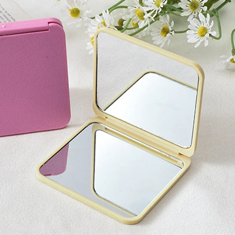 A pink portable makeup mirror with a clear double-sided reflective surface is shown next to a white floral arrangement.