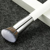 A silver-tipped makeup brush with a white handle rests on a dark gray textured surface.