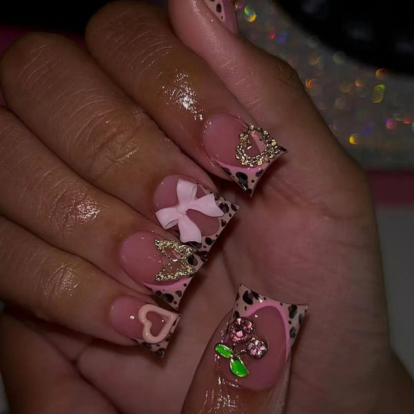 A hand displaying nude pink press-on nails decorated with leopard print and 3D gold charms.