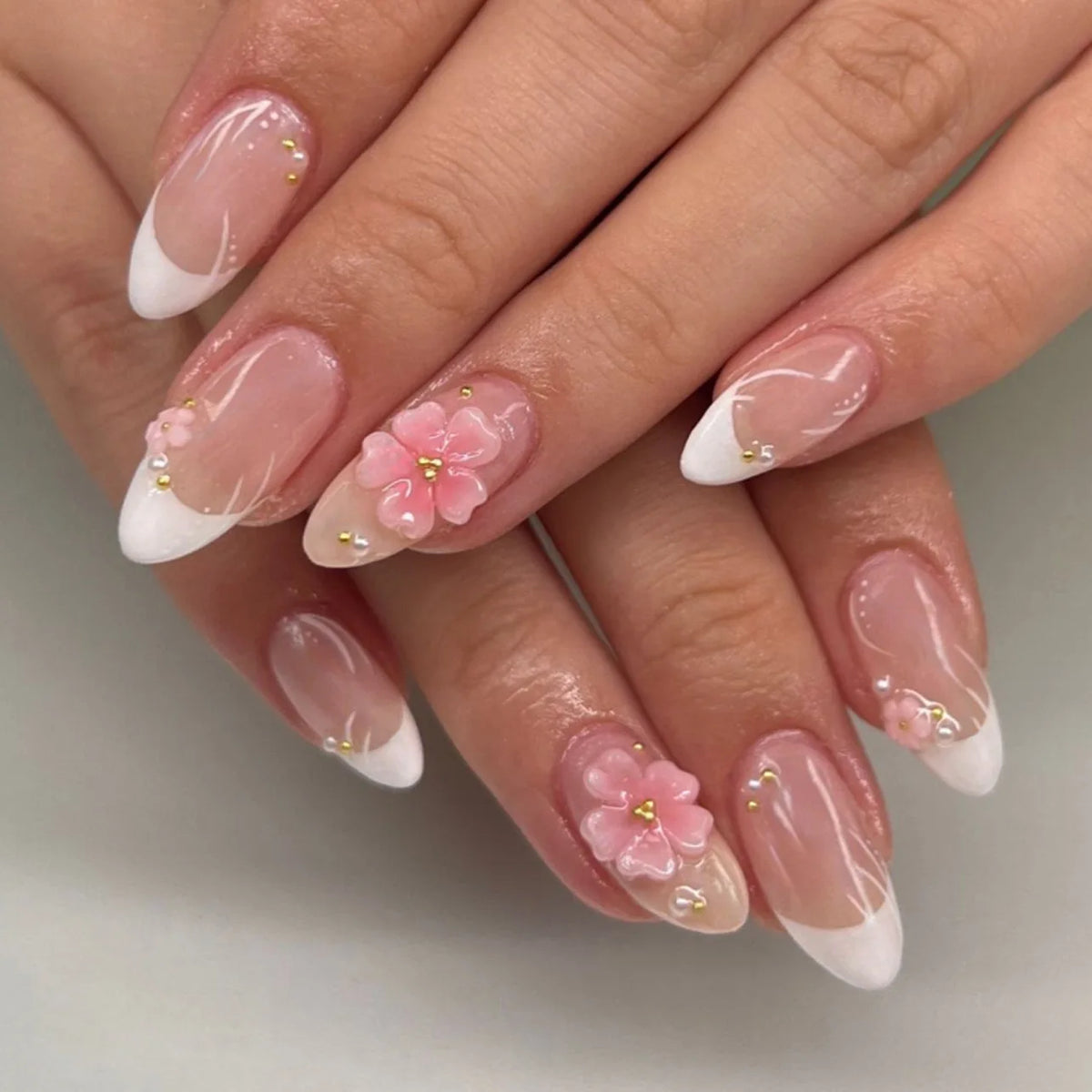 Gros plan d'une main montrant des kits de vernis press-on en forme de fleur d'amande avec des accents rose et blanc.