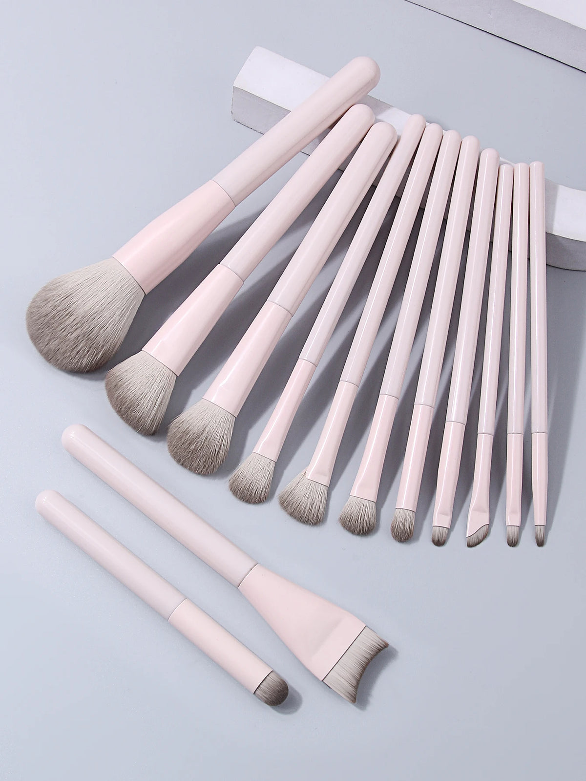 A set of thirteen professional makeup brushes with light pink handles and grey bristles arranged on a white surface.