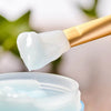 A silicone facial mask applicator brush with a gold handle is shown applying a white cream to a jar.
