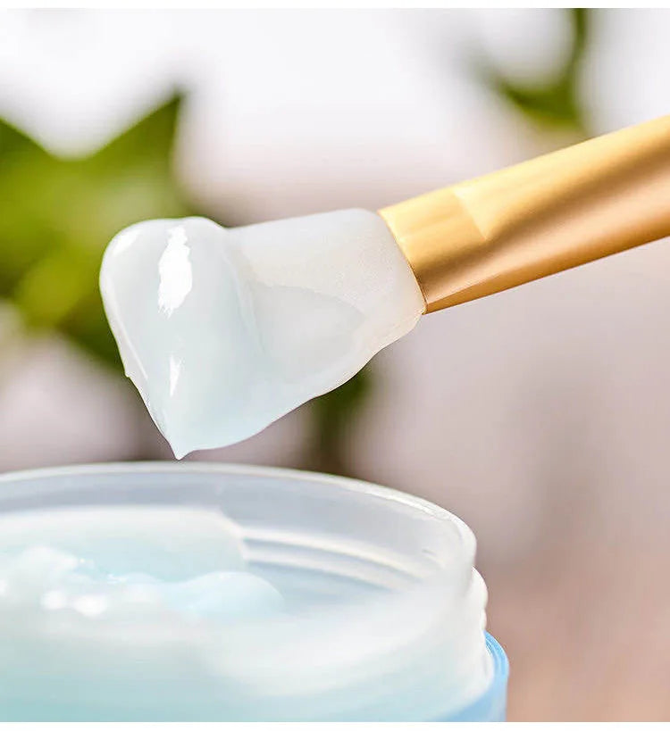 A silicone facial mask applicator brush with a gold handle is shown applying a white cream to a jar.