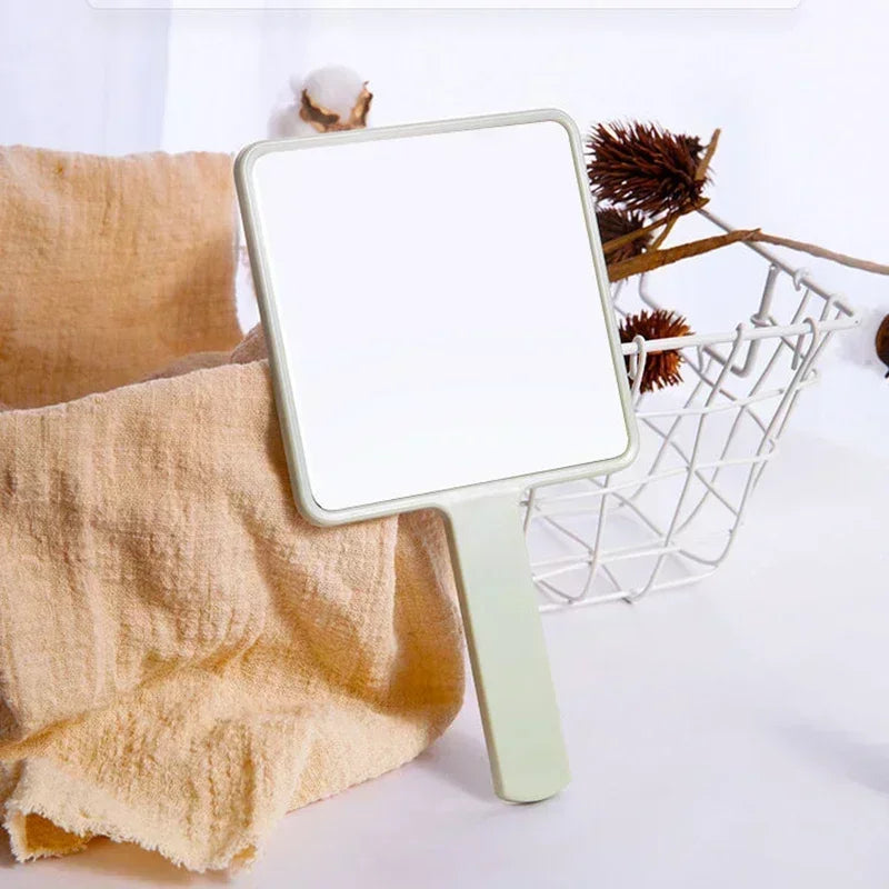 Un miroir de maquillage rectangulaire vert clair, tenu à la main, est exposé à côté d'une serviette beige et d'un panier déco