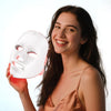 Una mujer joven sonriendo mientras sostiene una mascarilla facial translúcida con luces LED rojas para rejuvenecimiento de la