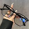 A hand holds a pair of black minimalist square reading glasses against a blurred gray background.
