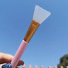 A pink silicone facial mask applicator brush with a white silicone pad is held against a clear blue sky.