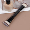 A black double-ended makeup brush with a fluffy pink and white synthetic hair is shown on a light surface.