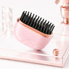 A pink portable hair straightener with black bristles rests on a metallic tray.