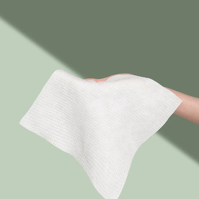 A hand is holding a white, textured disposable face towel against a muted green background.