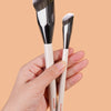 A hand holds a set of professional makeup brushes with silver ferrules and white handles against a beige background.