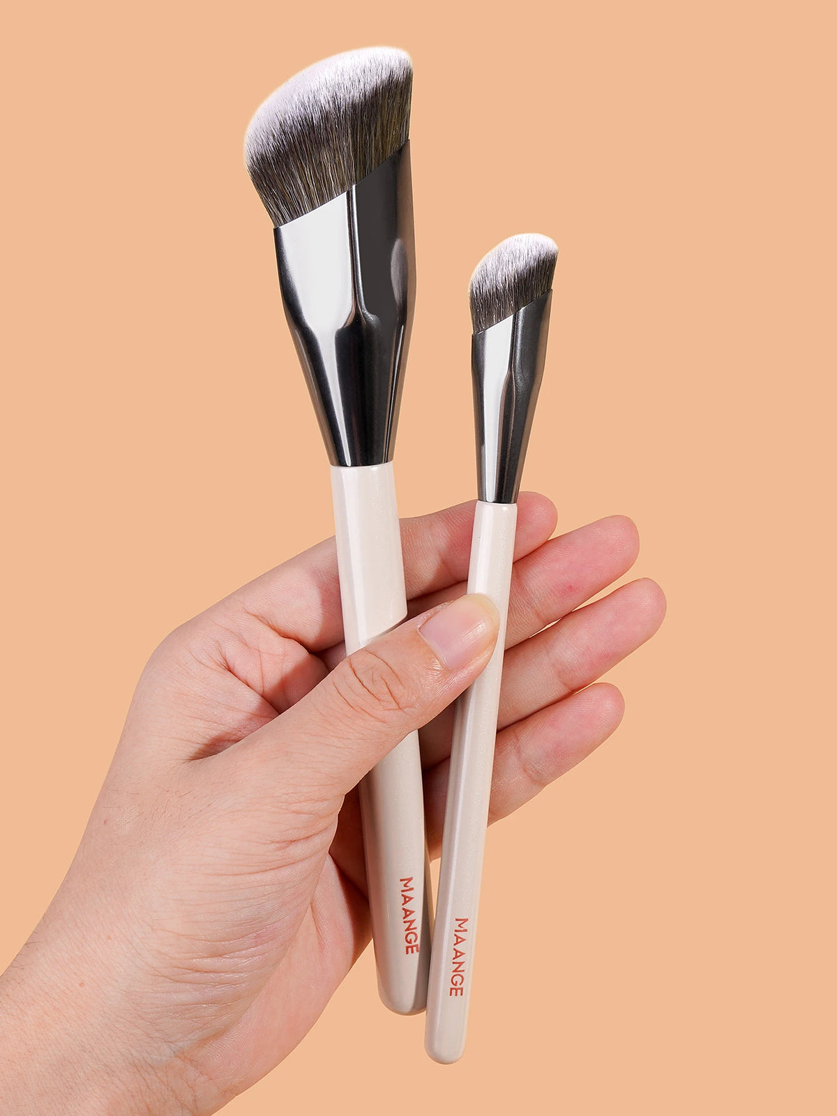 A hand holds a set of professional makeup brushes with silver ferrules and white handles against a beige background.