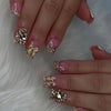 A close-up of manicured nails featuring nude pink press-on nails decorated with gold leopard print and 3D charms.