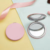 A pink portable magnifying makeup mirror is shown open on a light blue surface next to a yellow background.