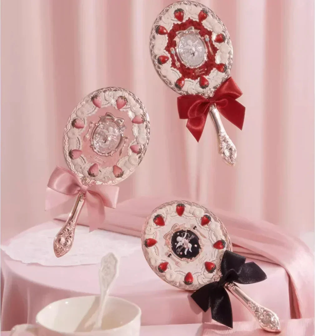 A set of decorative hand holding makeup mirrors with red bows and silver accents displayed on pink fabric.