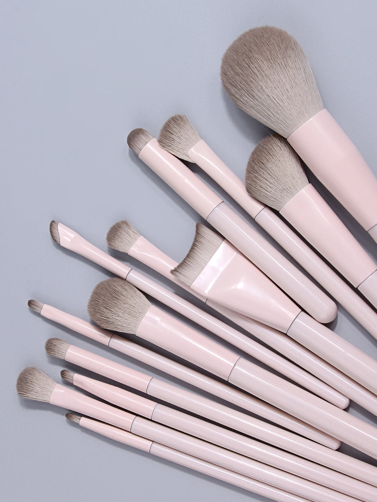A collection of thirteen professional makeup brushes with light pink handles and natural hair bristles.