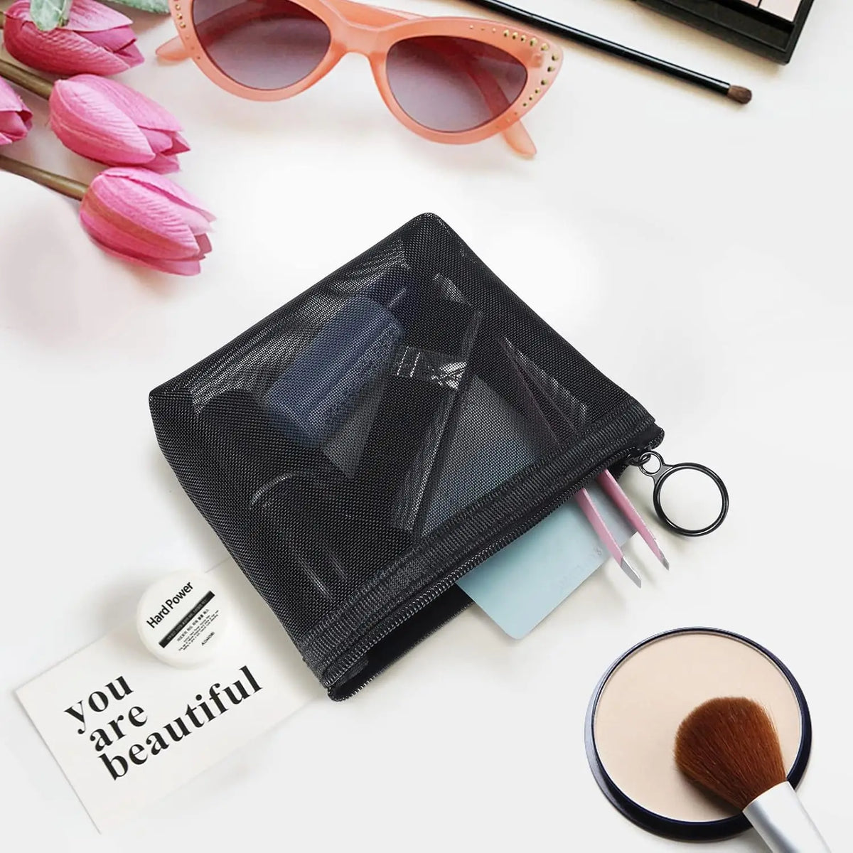Un sac de maquillage noir en filet à côté de tulipes roses, des lunettes de soleil et un pinceau de maquillage.