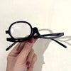 A hand holds a pair of black adjustable magnifying reading glasses with clear lenses against a light background.