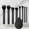 A set of nine black makeup brushes displayed vertically on a white surface with a large black brush head in the foreground.